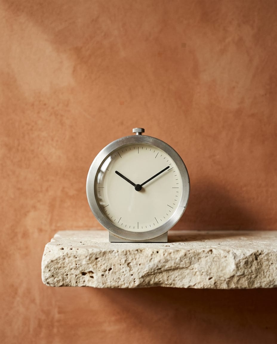 A table clock with a white dial in a brushed aluminium case on a limestone ledge against a terracotta wall